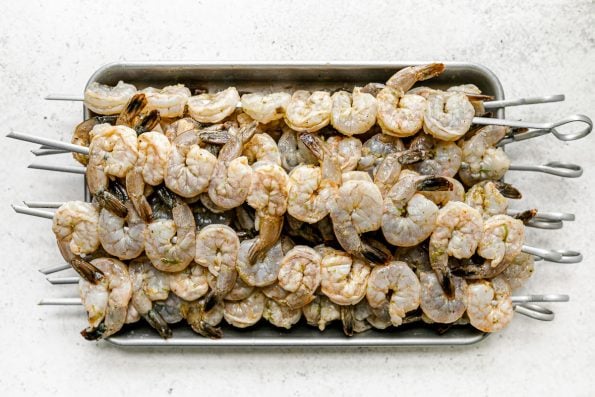 Multiple skewers of zesty marinated jumbo shrimp are stacked on top of an aluminum baking sheet. The baking sheet sits on top of a white & light gray textured surface.