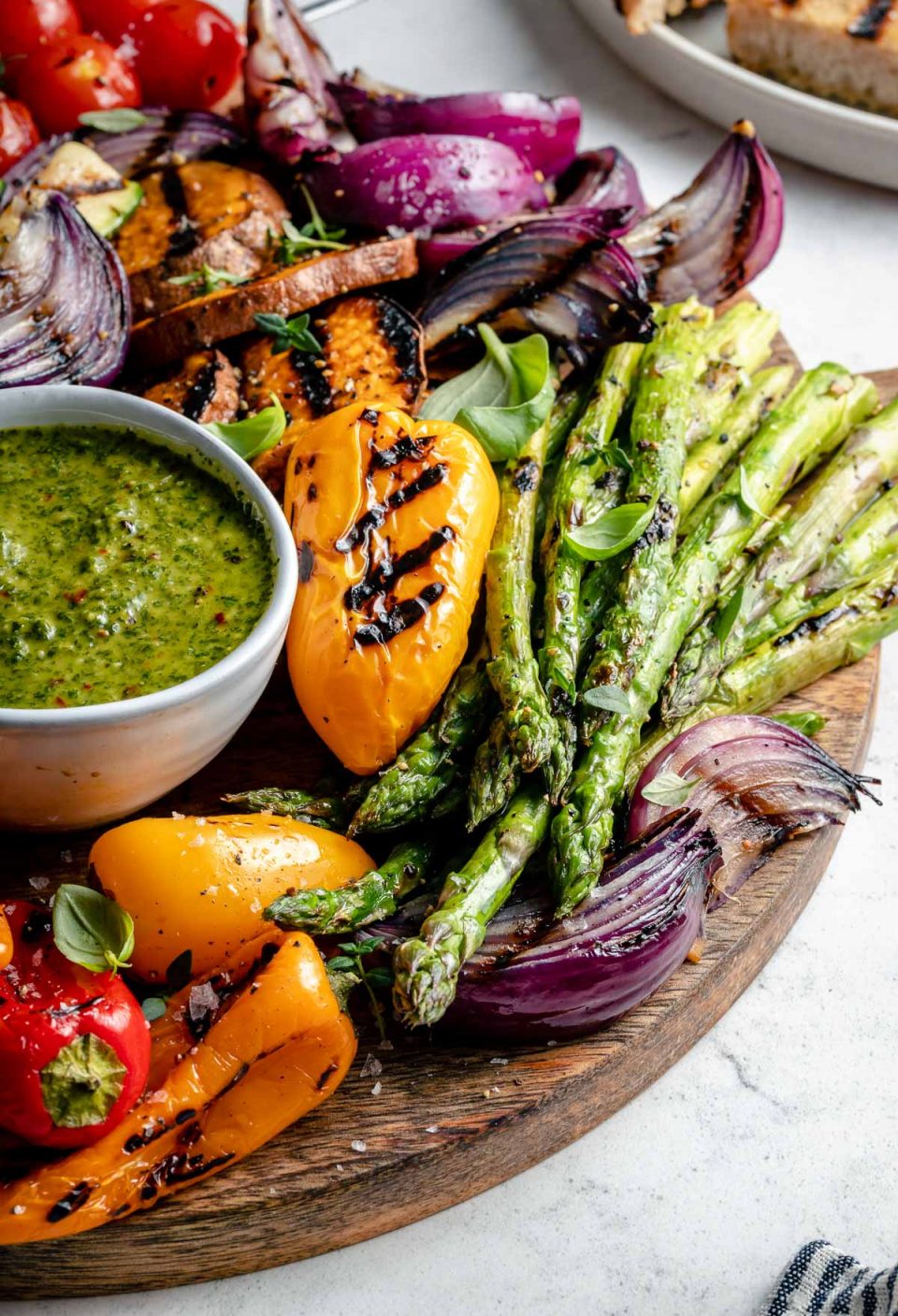 An angled shot of grilled veggies arranged on top of a round wooden cutting board. The grilled veggies include grilled onion, grilled tomato skewers, grilled asparagus, grilled sweet potatoes, & grilled bell peppers. The grilled veggies are garnished with fresh herbs & surround a small white bowl of chimichurri sauce for dipping that also rests on the cutting board. A white ceramic plate filled with grilled bread sits in the background and a small portion of a blue & white linen napkin rests in the foreground. All serving ware sits on top of a blue & white marbled surface.