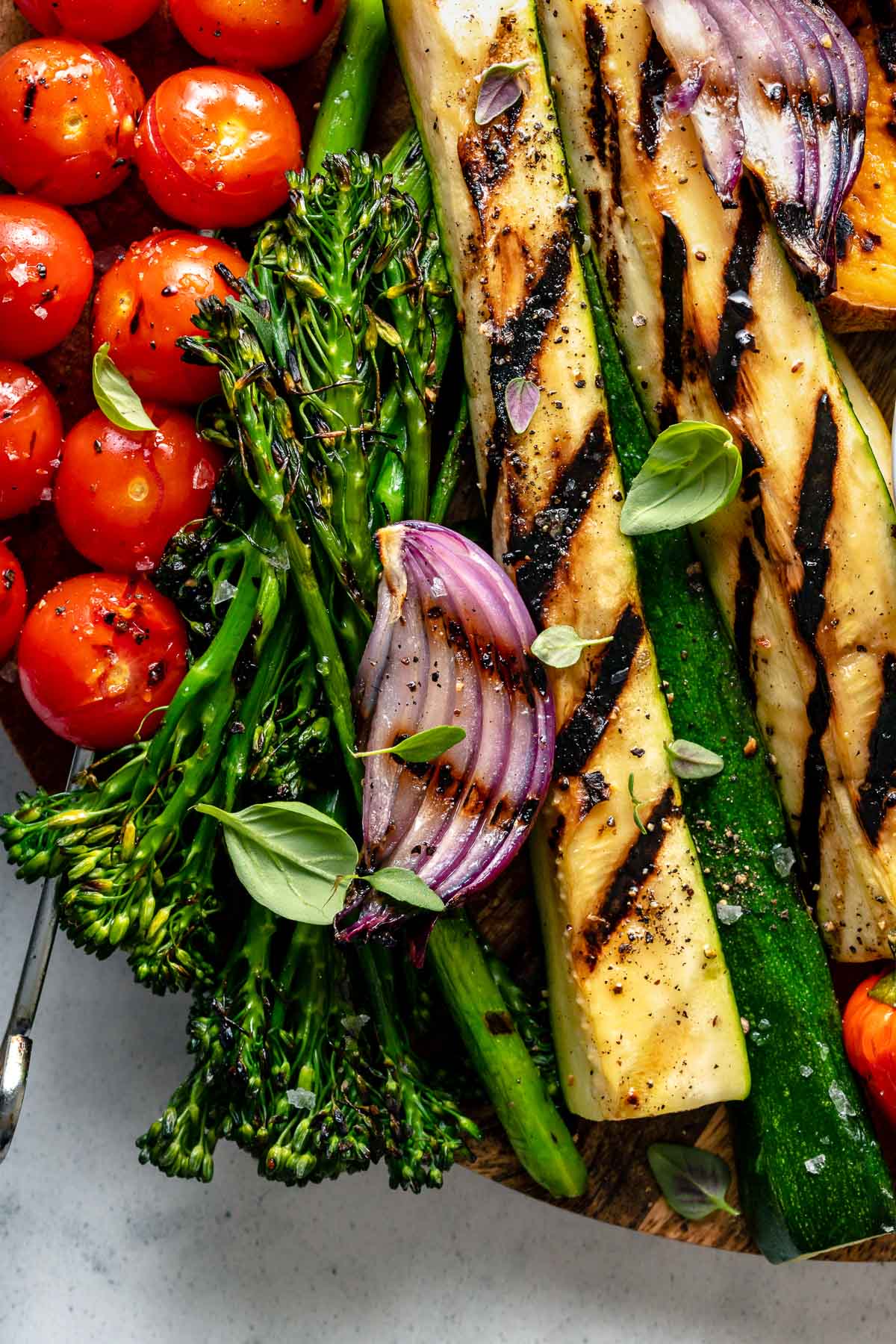 A close up for grilled veggies arranged on top of a round wooden cutting board. The close up includes grilled onion, grilled tomato skewers, grilled zucchini, & grilled broccolini garnished with fresh herbs. The wooden cutting board sits on top of a blue & white marbled surface.