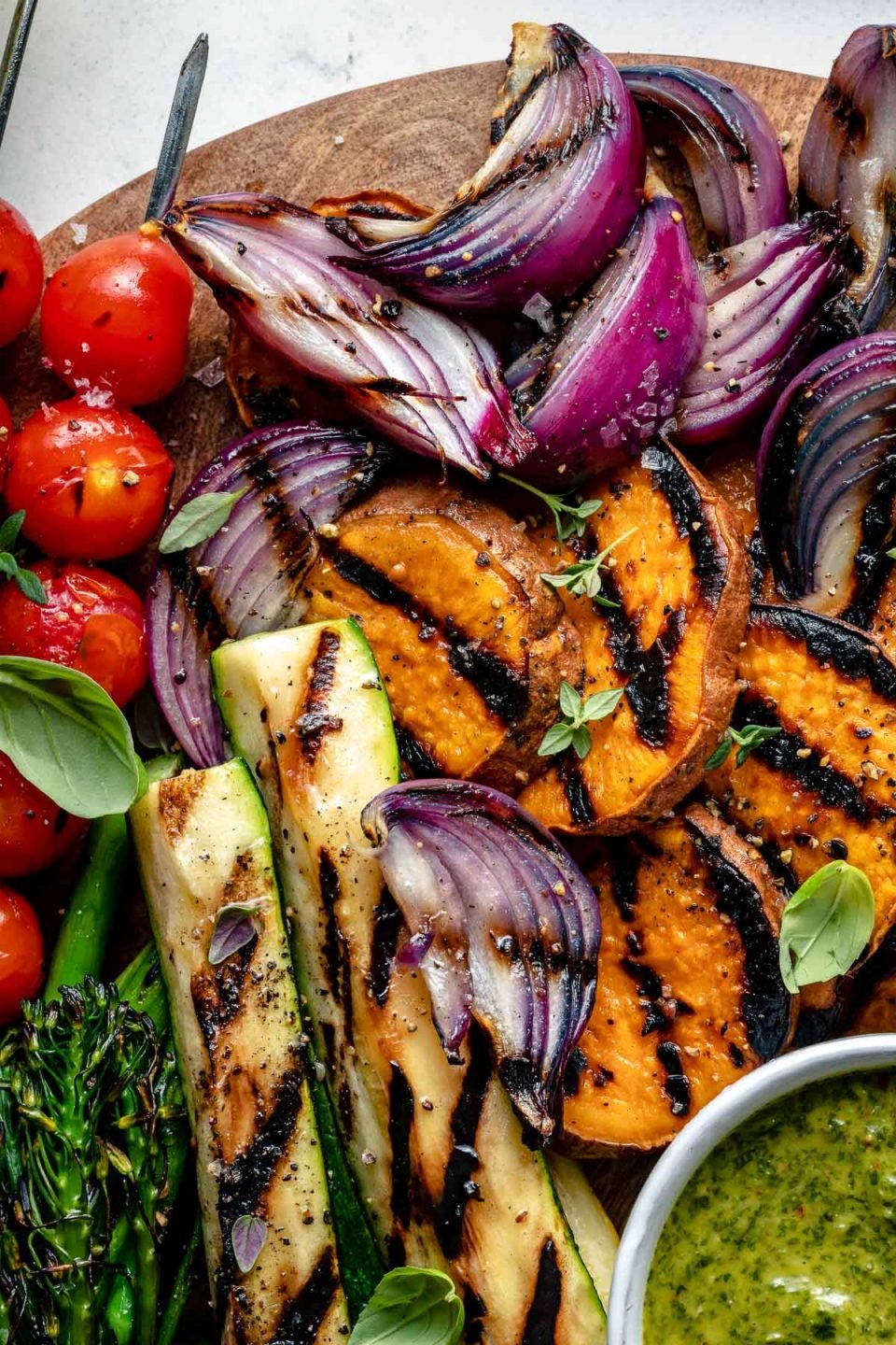 A close up for grilled veggies arranged on top of a round wooden cutting board. The close up includes grilled onion, grilled sweet potatoes, grilled tomato skewers, grilled zucchini, & grilled broccolini garnished with fresh herbs. The veggies are arranged around a small white bowl of chimichurri sauce for dipping that also sits on top of the cutting board. The wooden cutting board sits on top of a blue & white marbled surface.