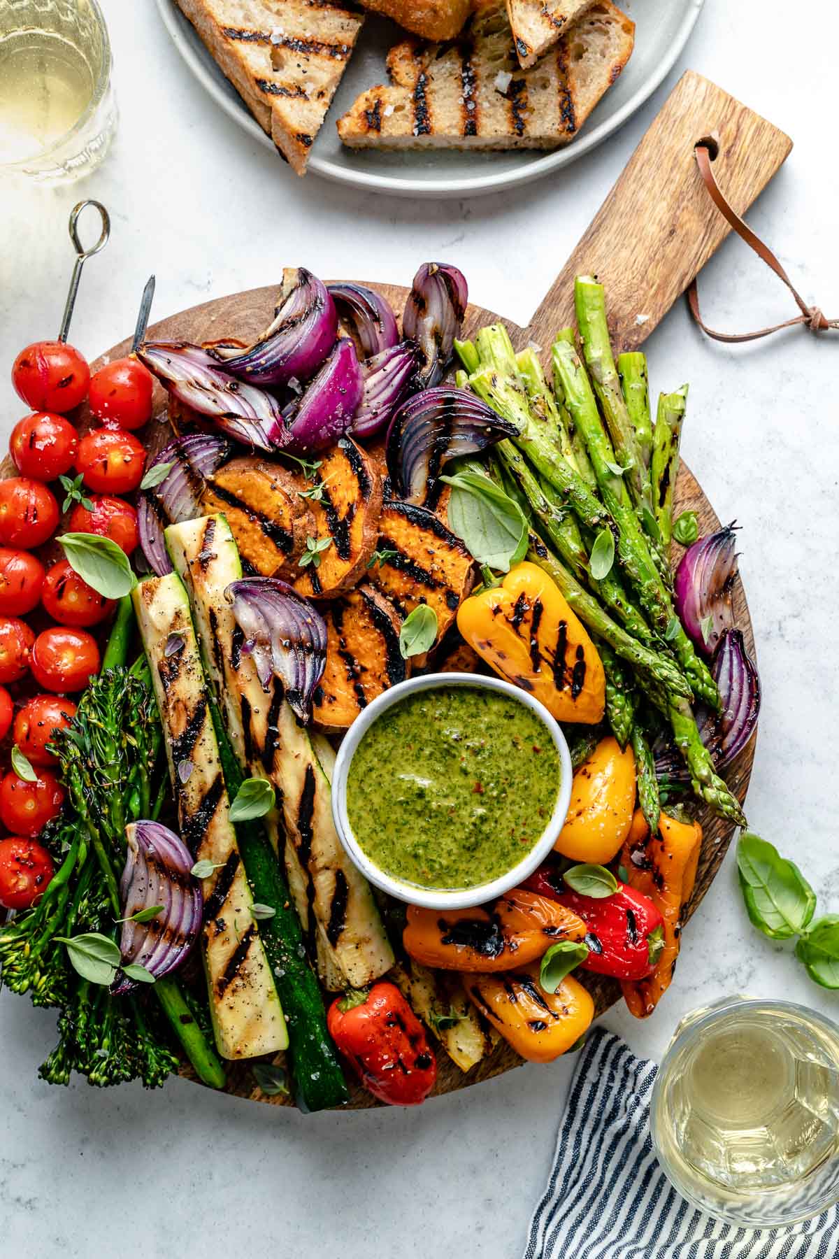 Grilled veggies including grilled cherry tomato skewers, grilled broccolini, grilled zucchini spears, grilled onion, grilled bell peppers, & grilled asparagus are arranged on a round wooden cutting board for serving. The grilled veggies are garnished with fresh herbs & surround a small white bowl of chimichurri sauce for dipping that also rests on the cutting board. A white ceramic plate filled with grilled bread, two small glasses of wine, fresh basil & a blue & white linen napkin surround the cutting board. All serving ware sits on top of a blue & white marbled surface.