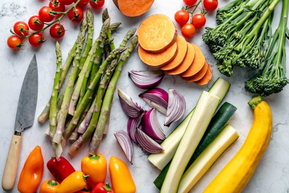 Grilled vegetable platter ingredients arranged on a white & blue-gray marble surface - cherry tomatoes, sweet potatoes, broccolini, asparagus, red onion, zucchini, yellow squash, & sweet peppers. A pairing knife rests on the surface next to the ingredients.