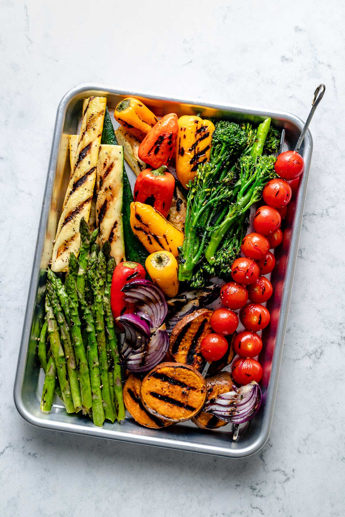 Grilled vegetables, including grilled tomato skewers, grilled red onion, grilled sweet potato, grilled broccolini, grilled bell peppers, grilled zucchini, & grilled asparagus seasoned with avocado oil, kosher salt, & ground black pepper are arranged on an aluminum baking sheet. The baking sheet sits on top of a blue & white marble surface.