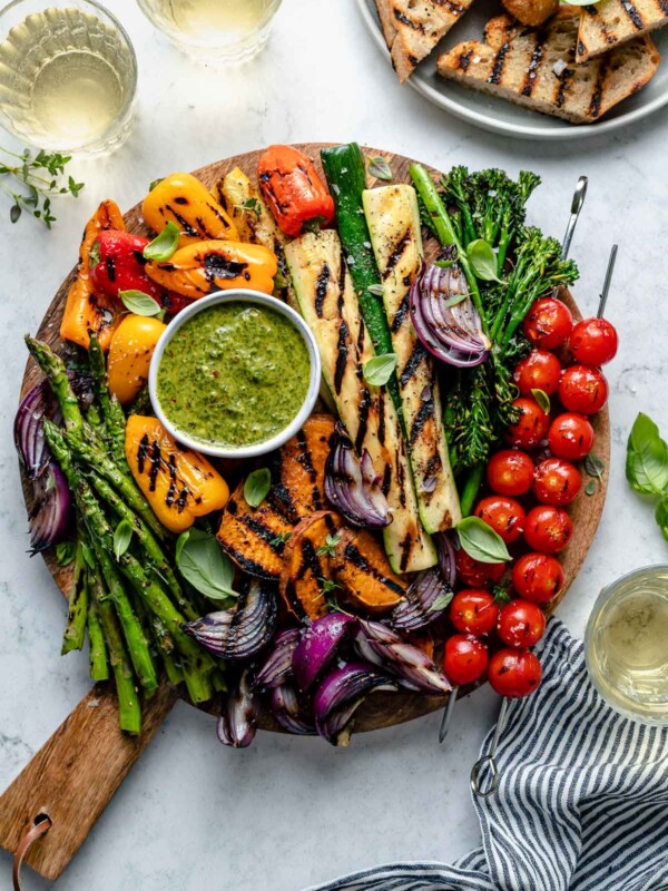 Grilled veggies including grilled cherry tomato skewers, grilled broccolini, grilled zucchini spears, grilled onion, grilled bell peppers, grilled sweet potatoes, & grilled asparagus are arranged on a round wooden cutting board for serving. The grilled veggies are garnished with fresh herbs & surround a small white bowl of chimichurri sauce for dipping that also rests on the cutting board. A white ceramic plate filled with grilled bread, three small glasses of white wine, fresh basil & herbs, a blue & white linen napkin surround the cutting board. All serving ware sits on top of a blue & white marbled surface.