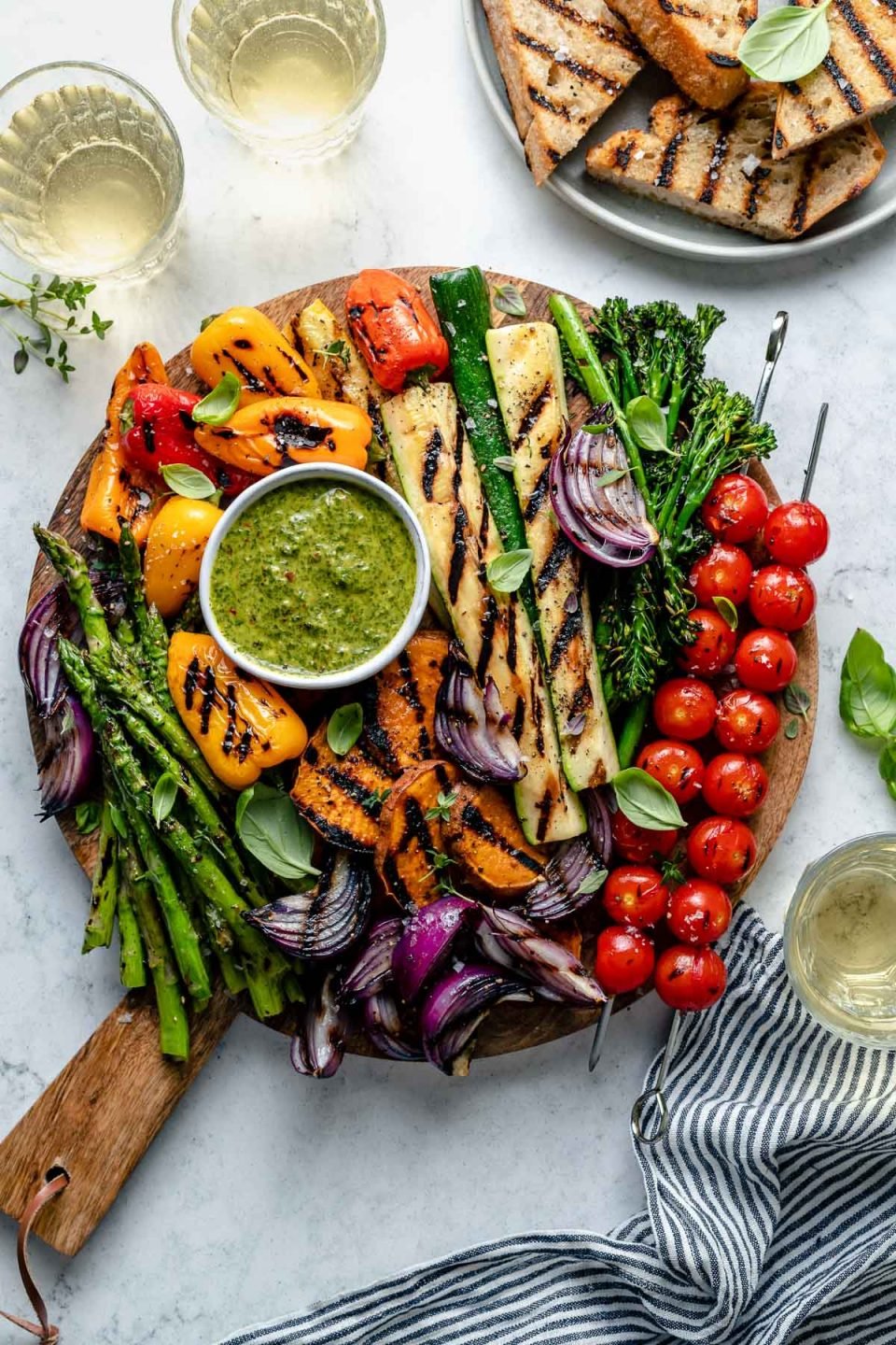 Grilled veggies including grilled cherry tomato skewers, grilled broccolini, grilled zucchini spears, grilled onion, grilled bell peppers, grilled sweet potatoes, & grilled asparagus are arranged on a round wooden cutting board for serving. The grilled veggies are garnished with fresh herbs & surround a small white bowl of chimichurri sauce for dipping that also rests on the cutting board. A white ceramic plate filled with grilled bread, three small glasses of white wine, fresh basil & herbs, a blue & white linen napkin surround the cutting board. All serving ware sits on top of a blue & white marbled surface.