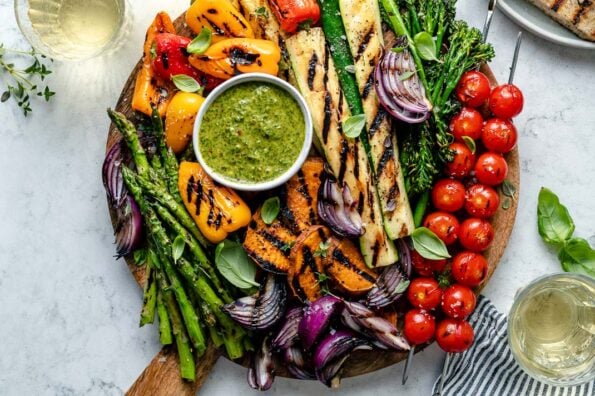 Grilled veggies including grilled cherry tomato skewers, grilled broccolini, grilled zucchini spears, grilled onion, grilled bell peppers, grilled sweet potatoes, & grilled asparagus are arranged on a round wooden cutting board for serving. The grilled veggies are garnished with fresh herbs & surround a small white bowl of chimichurri sauce for dipping that also rests on the cutting board. A white ceramic plate filled with grilled bread, three small glasses of white wine, fresh basil & herbs, a blue & white linen napkin surround the cutting board. All serving ware sits on top of a blue & white marbled surface.