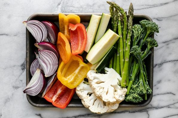 A variety of veggies including broccolini, asparagus, cauliflower, zucchini, onion, & peppers on a aluminum baking sheet. The baking sheet rests on a white & gray marble surface.