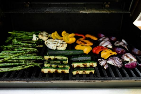 A straight on shot of a variety of grilled veggies including grilled broccolini, grilled asparagus, grilled cauliflower, grilled zucchini, grilled onion, & grilled peppers that are being grilled on gas grill grates. Many of the veggies have grill marks on them after being flipped to the other side.