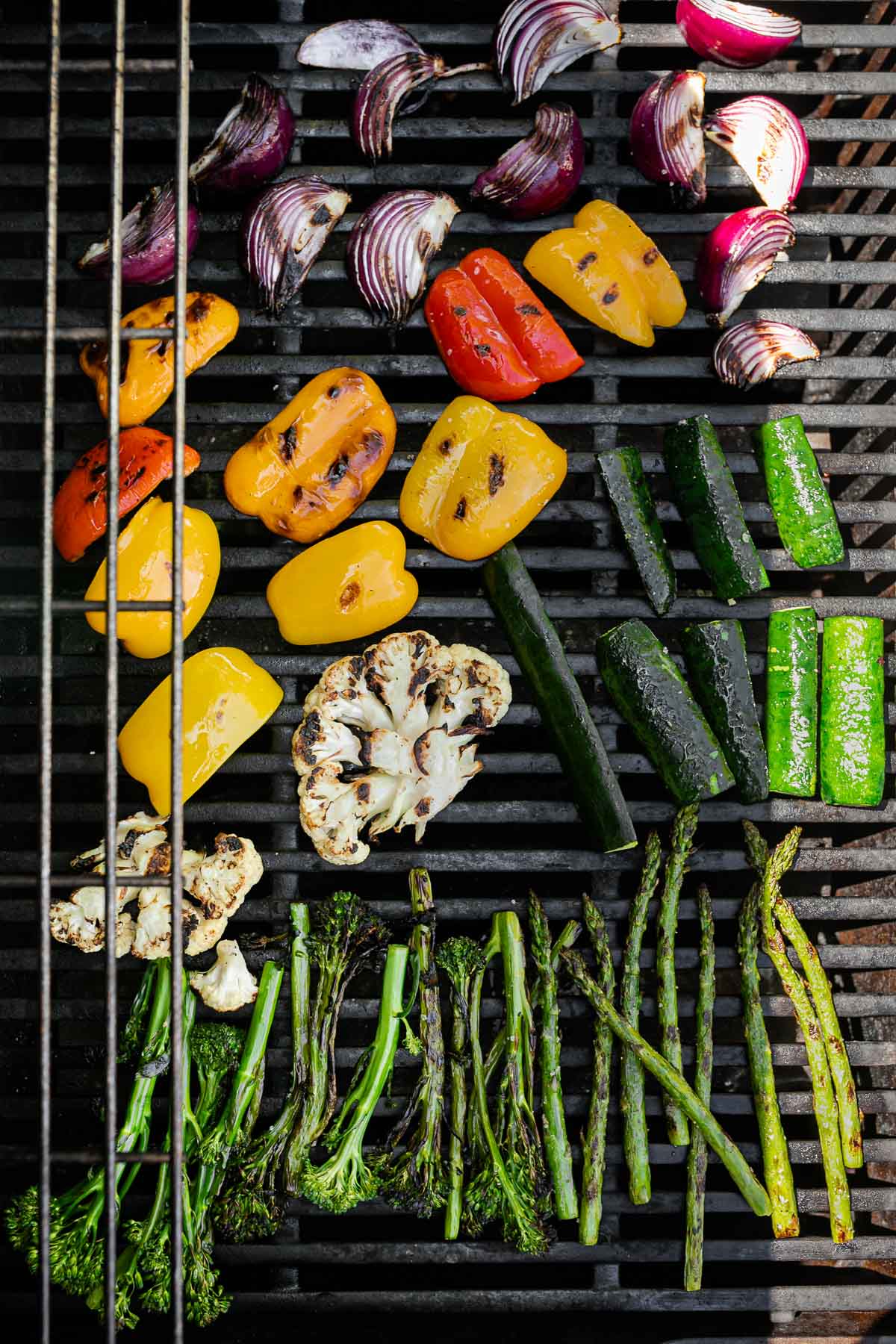 An overhead shot of a variety of grilled veggies including grilled broccolini, grilled asparagus, grilled cauliflower, grilled zucchini, grilled onion, & grilled peppers are being grilled on a gas grill grates. Many of the veggies have grill marks on them after being flipped to the other side.