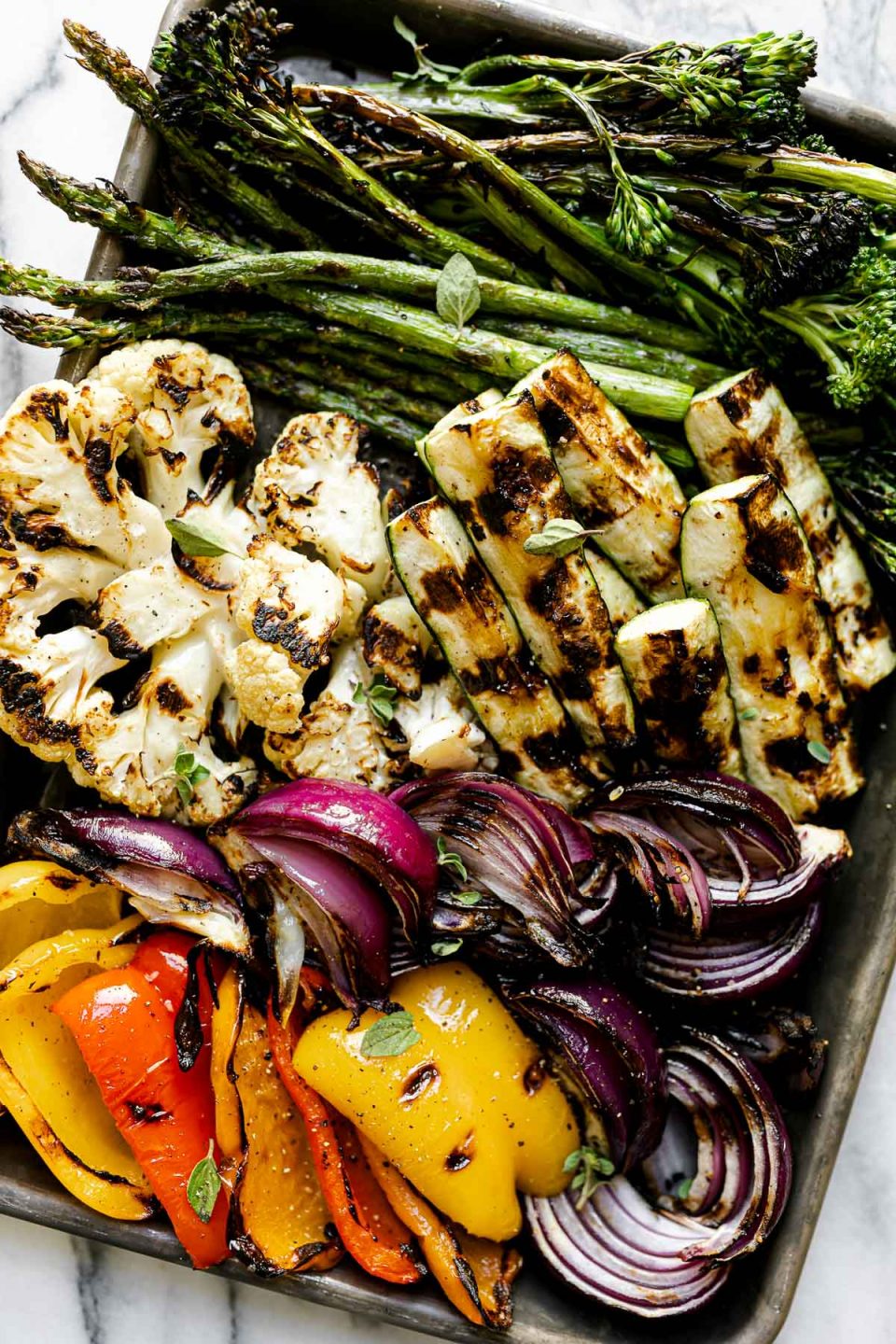 A variety of grilled veggies including grilled broccolini, grilled asparagus, grilled cauliflower, grilled zucchini, grilled onion, & grilled peppers on a aluminum baking sheet. The baking sheet rests on a white & gray marble surface.