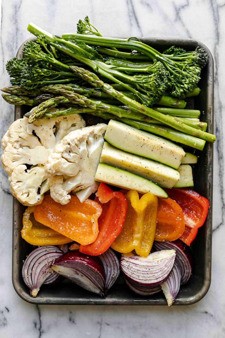 A variety of veggies including broccolini, asparagus, cauliflower, zucchini, onion, & peppers on a aluminum baking sheet. The baking sheet rests on a white & gray marble surface.