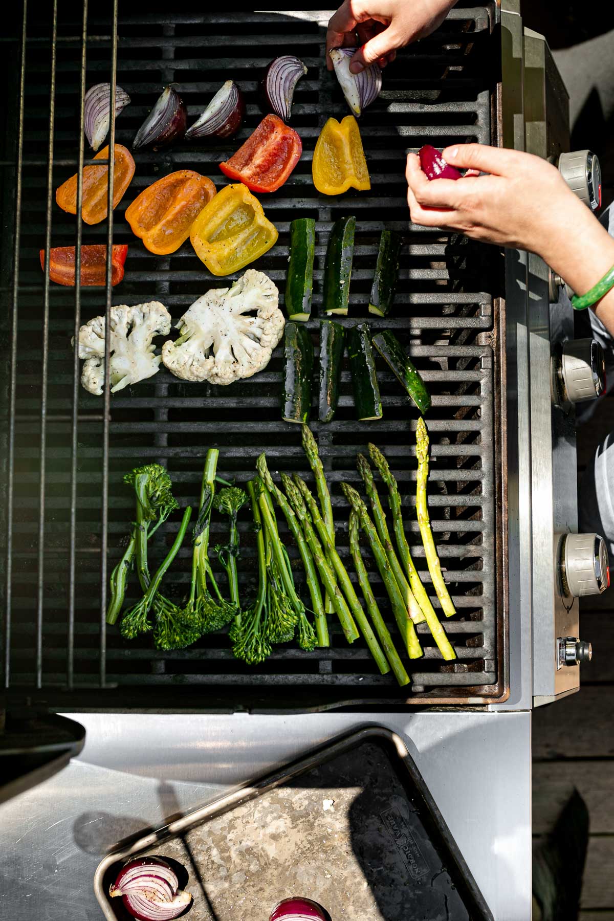 An overhead shot of a woman's hands placing a variety of raw veggies on top of gas grill grates for grilling. These veggies include broccolini, asparagus, cauliflower, zucchini, onion, & peppers. A aluminum baking sheet sits on the shelf to the left of the grill with additional onions ready to be placed on the grates.