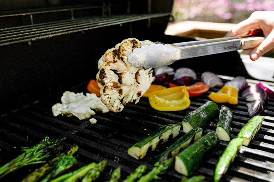 An angled shot of a variety of grilled veggies including grilled broccolini, grilled asparagus, grilled cauliflower, grilled zucchini, grilled onion, & grilled peppers that are being grilled on gas grill grates. A woman's hand holds a pair of tongs that is picking up a grilled cauliflower steak ready to flip it over. Many of the veggies have grill marks on them after being flipped to the other side.
