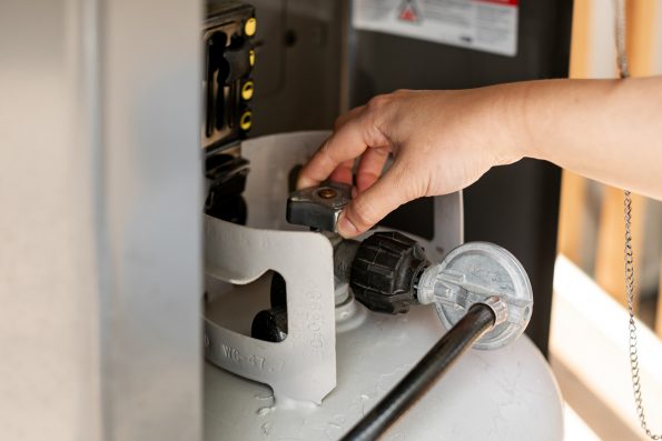 How to Start a Propane Grill, Step 1: A woman's hand reaches to a propane tank attached to a gas grill, turning it to open the valve.