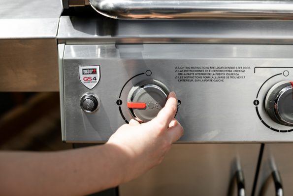 How to Start a Propane Grill, Step 2: A woman's hand reaches to open a burner valve of a Weber Genesis II propane grill.