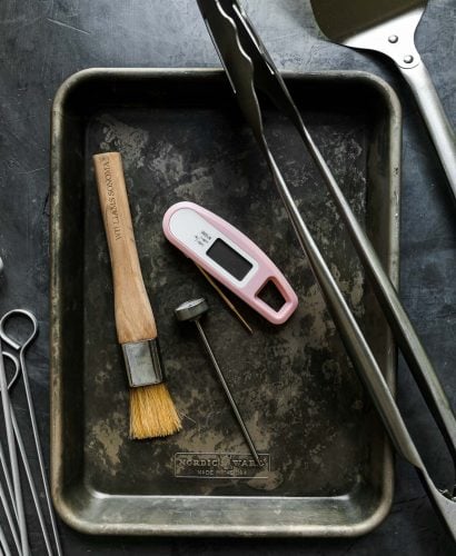 Grill tools arranged on a black surface: quarter sheet pans, stainless steel skewers, oven thermometer, instant read thermometer, pastry brush, grill tongs, grill spatula, grill brush.