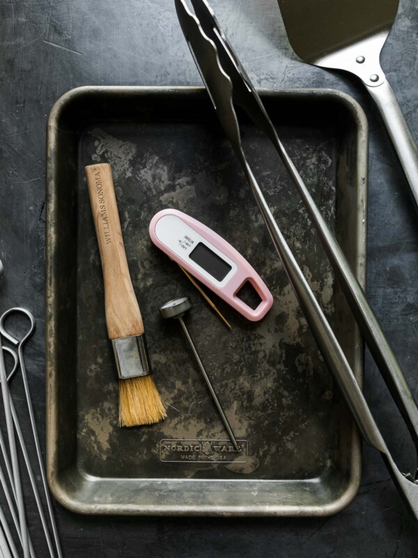 Grill tools arranged on a black surface: quarter sheet pans, stainless steel skewers, oven thermometer, instant read thermometer, pastry brush, grill tongs, grill spatula, grill brush.