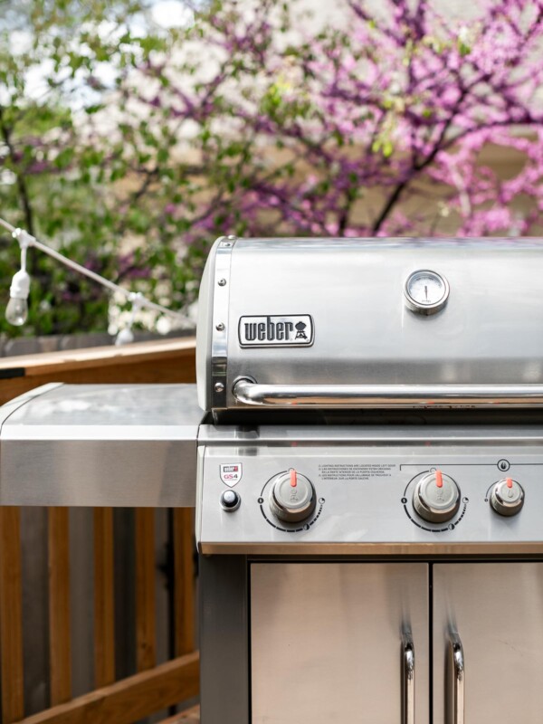 Weber Genesis II gas grill shown on deck. In the background, a blossoming tree and a house with beige siding.