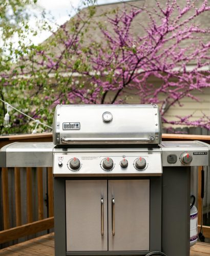Weber Genesis II gas grill shown on deck. In the background, a blossoming tree and a house with beige siding.