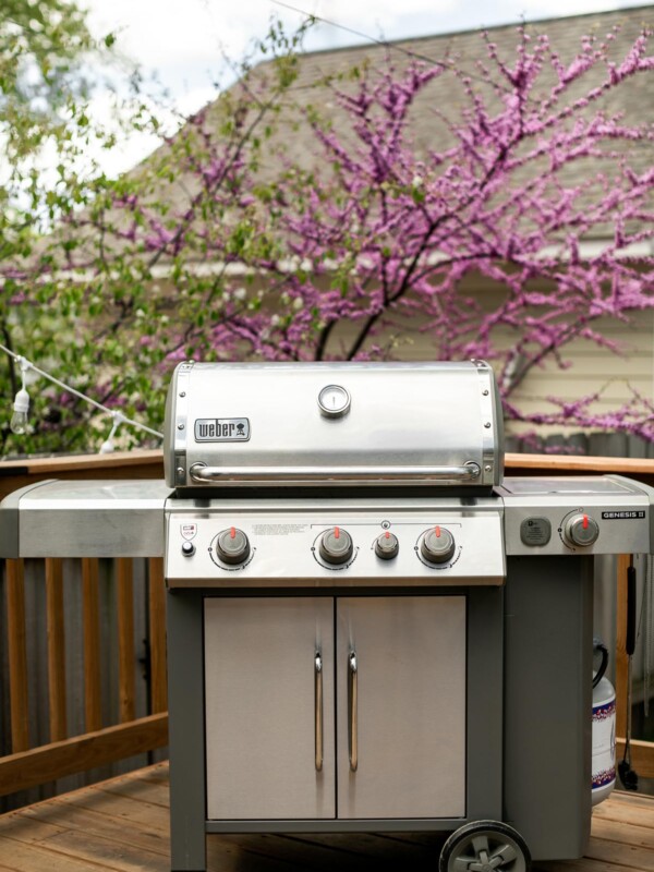 Weber Genesis II gas grill shown on deck. In the background, a blossoming tree and a house with beige siding.