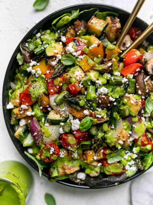 Chopped grilled vegetable salad in a large black serving bowl atop a gray linen napkin on a light gray surface. 2 gold serving spoons are nestled into the chopped vegetable salad. Fresh basil leaves & a jar of lemon basil vinaigrettes surround the serving bowl.