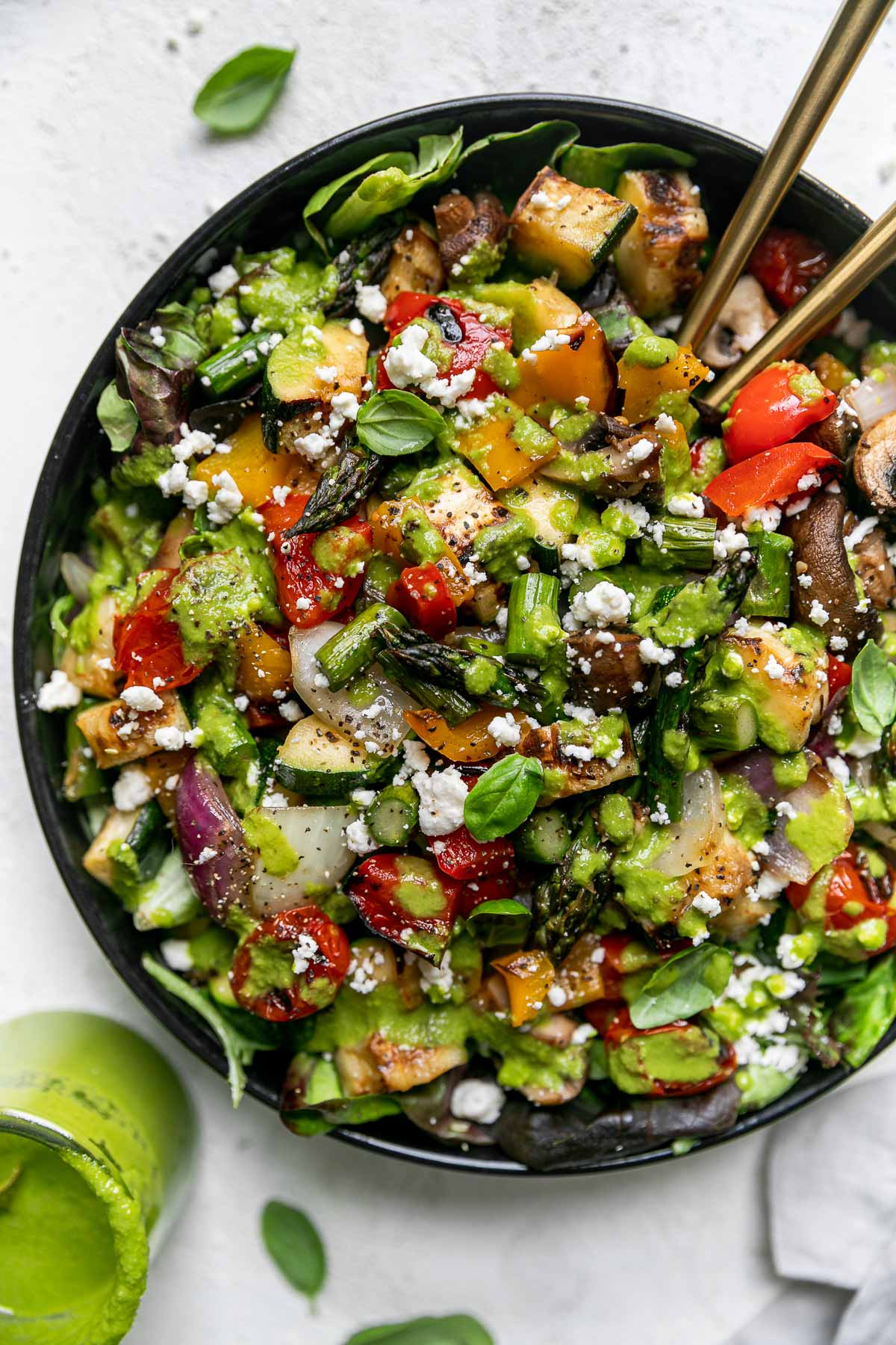 Chopped grilled vegetable salad in a large black serving bowl atop a gray linen napkin on a light gray surface. 2 gold serving spoons are nestled into the chopped vegetable salad. Fresh basil leaves & a jar of lemon basil vinaigrettes surround the serving bowl.