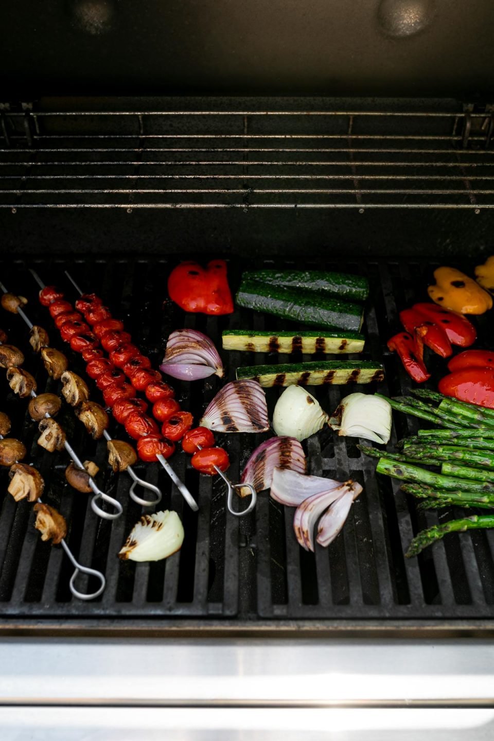 A straight on shot of a variety of grilled veggies including grilled asparagus, grilled zucchini, grilled onion, grilled bell peppers, grilled cherry tomatoes on skewers, & grilled mushrooms on skewers ​are being grilled on gas grill grates. Many of the veggies have grill marks on them after being flipped to the other side