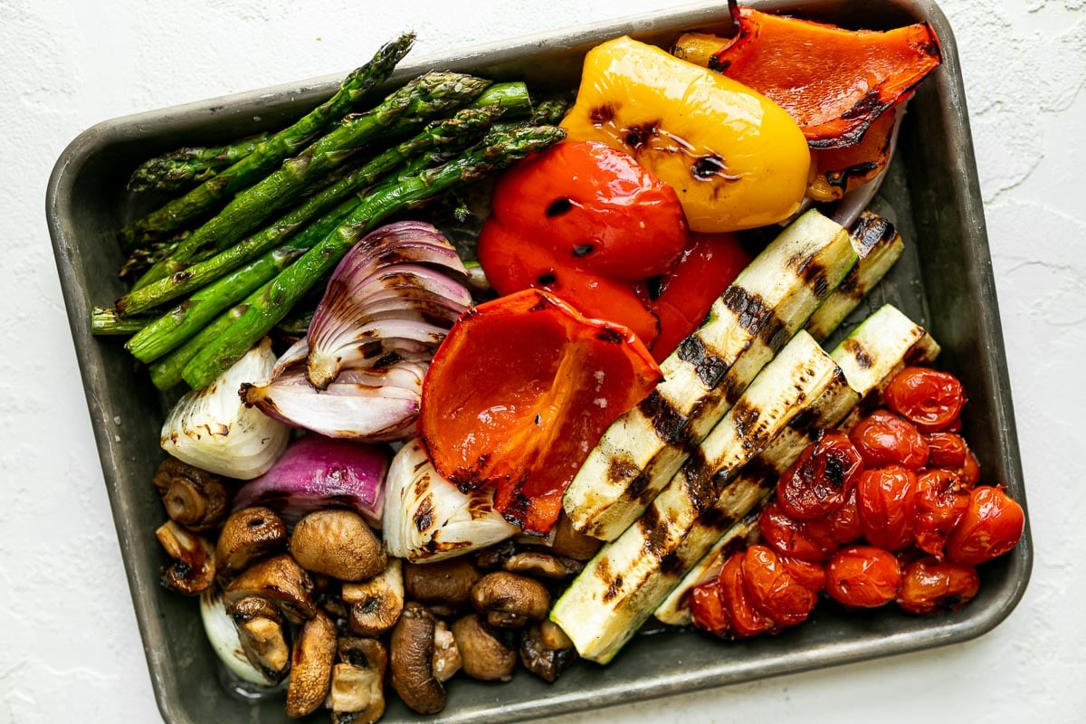 A variety of grilled veggies including grilled asparagus, grilled peppers, grilled onion, grilled mushrooms, grilled tomatoes, & grilled zucchini on an aluminum baking sheet. The baking sheet rests on a white surface.