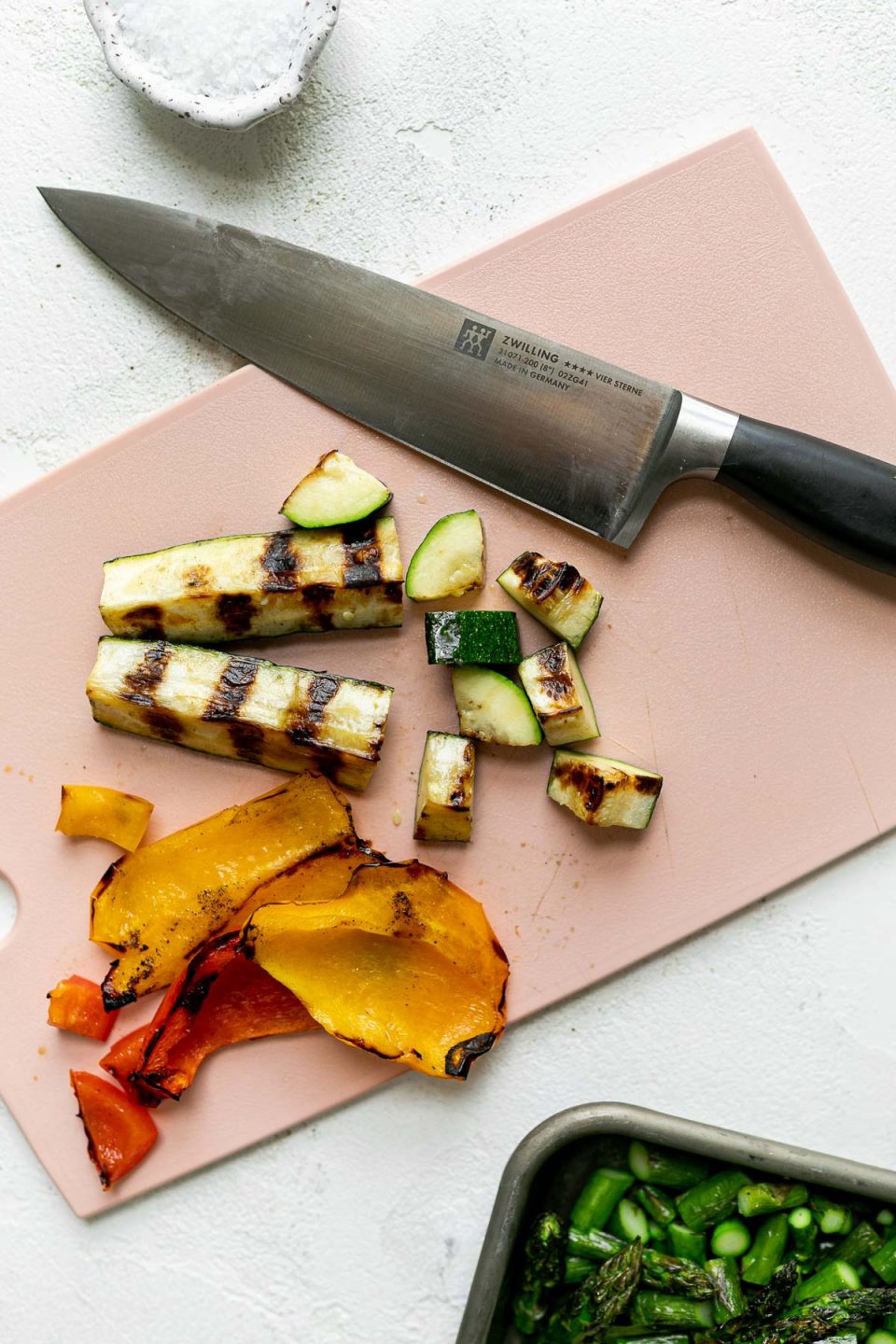 Sliced & diced grilled zucchini and bell pepper sit on top of a pink plastic cutting board a Zwilling knife used for chopping the veggies rests on top of the cutting board. The cutting board & knife sit atop a white surface. A small pinch bowl of kosher salt & a baking sheet full of grilled veggies surround the cutting board.