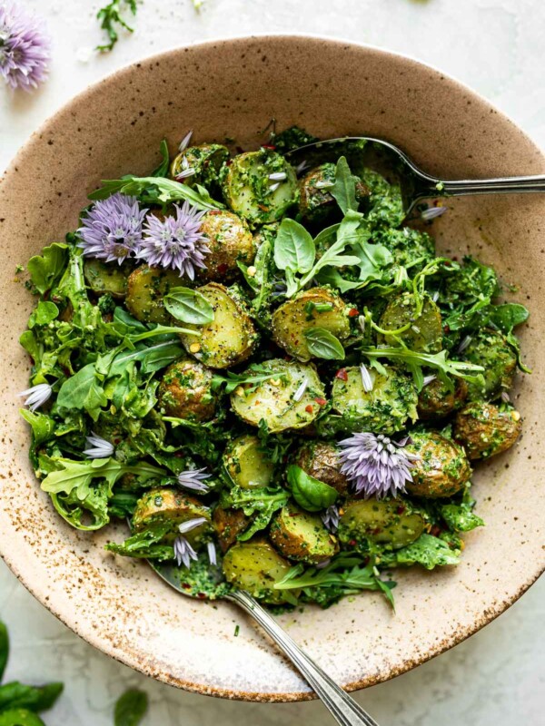 Green goddess pesto potato salad in ceramic serving bowl atop a white surface. The potato salad is tossed & topped with purple chive blossoms & has serving spoons nestled in it. The bowl is surrounded by fresh herbs, loose arugula leaves, & chive blossoms.