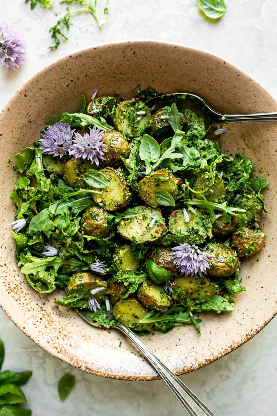 Green goddess pesto potato salad in ceramic serving bowl atop a white surface. The potato salad is tossed & topped with purple chive blossoms & has serving spoons nestled in it. The bowl is surrounded by fresh herbs, loose arugula leaves, & chive blossoms.