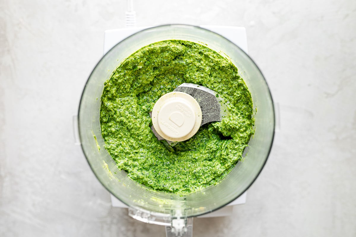 Blended green goddess pesto in a food processor carafe, atop a white surface.