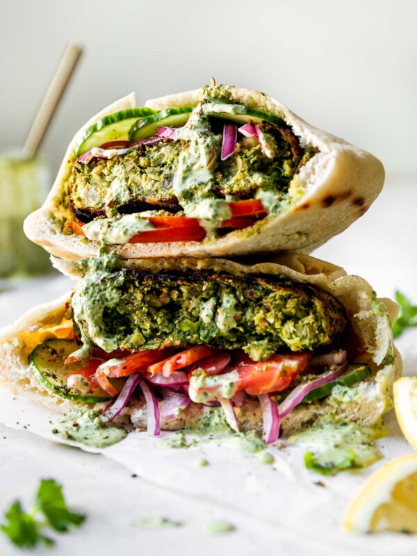 Two Grilled Falafel Burgers stacked on top of each other served inside a pita stuffed with a smear of hummus, thinly sliced tomatoes, cucumbers, & pickled red onion. The chickpea veggie burgers have then been drizzled with spicy green tahini sauce. The burgers are on a small piece of wrinkled parchment paper on top of a white surface, with a jar of tahini sauce and sliced lemons in the background & foreground.