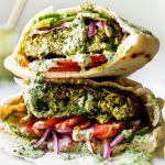 Two Grilled Falafel Burgers stacked on top of each other served inside a pita stuffed with a smear of hummus, thinly sliced tomatoes, cucumbers, & pickled red onion. The chickpea veggie burgers have then been drizzled with spicy green tahini sauce. The burgers are on a small piece of wrinkled parchment paper on top of a white surface, with a jar of tahini sauce and sliced lemons in the background & foreground.