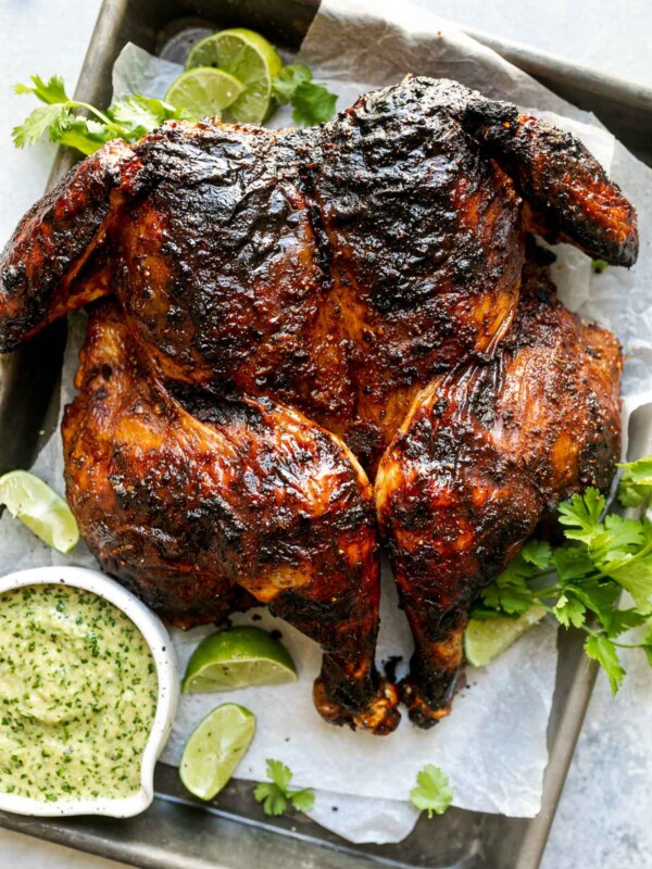 Grilled spatchcocked chicken with crispy, charred skin, sitting atop a parchment-lined baking sheet surrounded by lime wedges, fresh cilantro, & charred jalapeno sauce. The baking sheet sits atop a light blue surface, surrounded by fresh cilantro leaves.