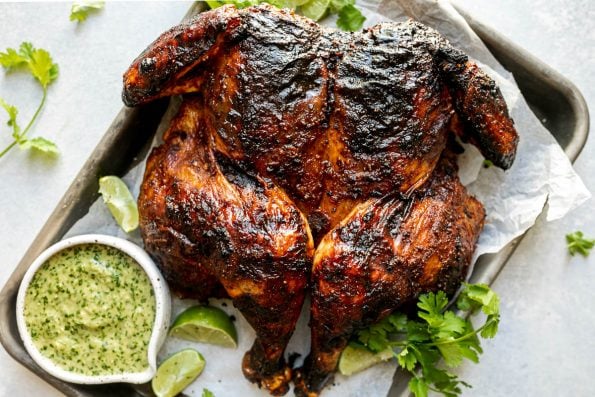 Grilled spatchcocked chicken with crispy, charred skin, sitting atop a parchment-lined baking sheet surrounded by lime wedges, fresh cilantro, & charred jalapeno sauce. The baking sheet sits atop a light blue surface, surrounded by fresh cilantro leaves.