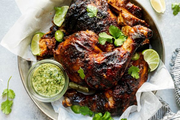 Butchered grilled whole chicken in a tin tray, topped with lime wedges, sliced jalapeno, & cilantro leaves. The tray sits atop a light blue surface, surrounded by fresh lime wedges, cilantro leaves, & a dark gray & white striped linen napkin.