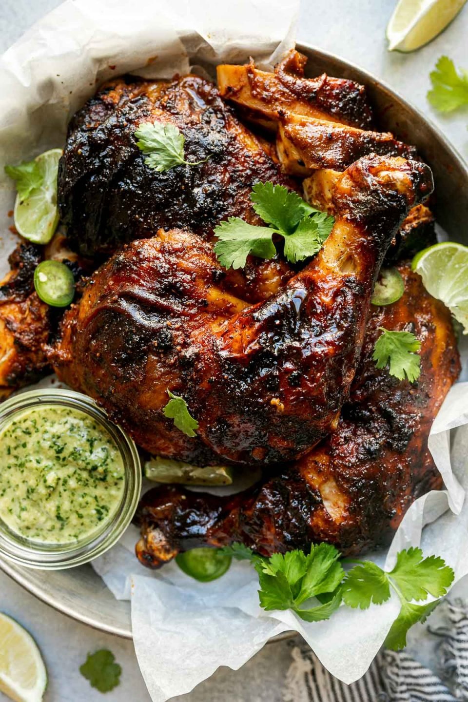 Butchered grilled whole chicken in a tin tray, topped with lime wedges, sliced jalapeno, & cilantro leaves. The tray sits atop a light blue surface, surrounded by fresh lime wedges, cilantro leaves, & a dark gray & white striped linen napkin.