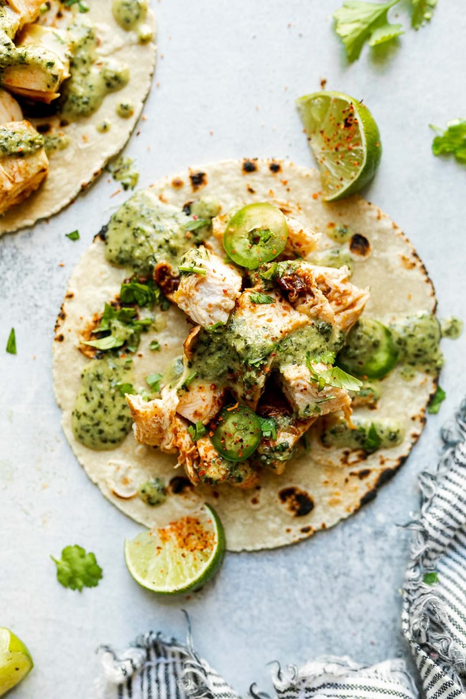 2 grilled chicken tacos, topped with creamy jalapeno sauce & sliced jalapeno. The tacos sit atop a light blue surface, surrounded by cilantro leaves, Tajin-dusted lime wedges, & a gray & white striped linen napkin.