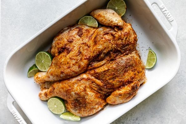 How to grill whole chicken, Step 3: Chili-lime wet rubbed chciken in a large ceramicbaking dish, surrounded by limes. The dish sits atop a light blue surface.