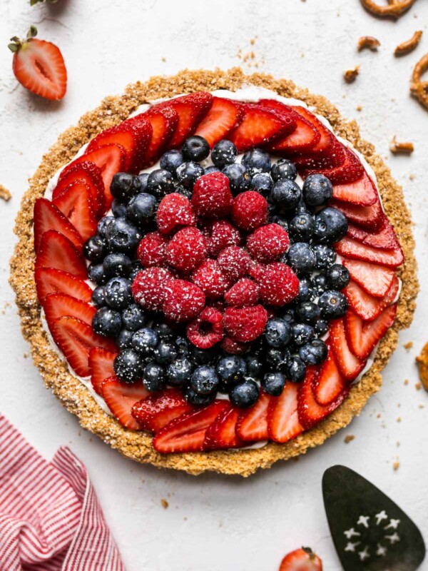 Close up of no-bake summer berry cheesecake tart with pretzel graham cracker crust. The tart is on a white textured surface, topped with fresh strawberries, blueberries, raspberries, & a sprinkle of powdered sugar. Surrounding the tart are loose berries, whole and crumbled pretzels, graham cracker crumbs, a white and red linen napkin, & a pie server utensil.