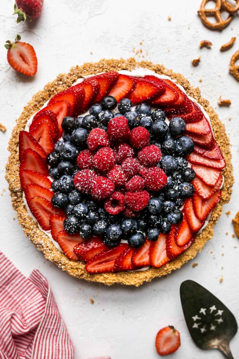 Close up of no-bake summer berry cheesecake tart with pretzel graham cracker crust. The tart is on a white textured surface, topped with fresh strawberries, blueberries, raspberries, & a sprinkle of powdered sugar. Surrounding the tart are loose berries, whole and crumbled pretzels, graham cracker crumbs, a white and red linen napkin, & a pie server utensil.