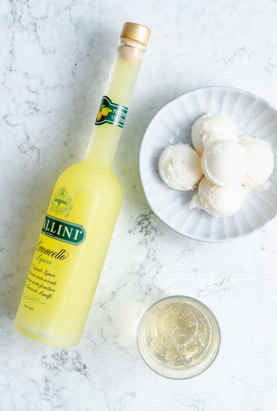 Limoncello, a bowl of sorbet and a glass of prosecco sit atop a white marbled surface.