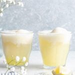 A side shot of two limoncello prosecco cocktails atop a white surface with a grey background. Sprigs of baby's breath flowers, a champagne cork, and lemon wedges are in the background.