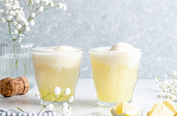 A side shot of two limoncello prosecco cocktails atop a white surface with a grey background. Sprigs of baby's breath flowers, a champagne cork, and lemon wedges are in the background.
