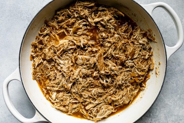 Shredded beer-braised chicken for green enchiladas in a ceramic braising pan atop a light blue surface.