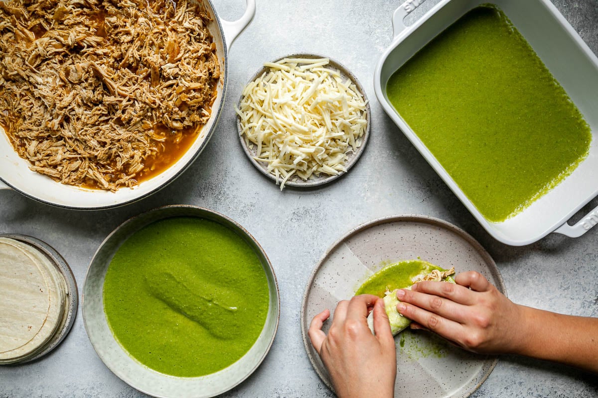 Green enchiladas assembly – green chicken enchiladas components (shredded chicken, shredded cheese, corn tortillas, & green enchiladas sauce atop a light blue surface. A woman's hands reach into the frame, wrapping chicken & cheese into a enchilada sauce-coated corn tortilla.