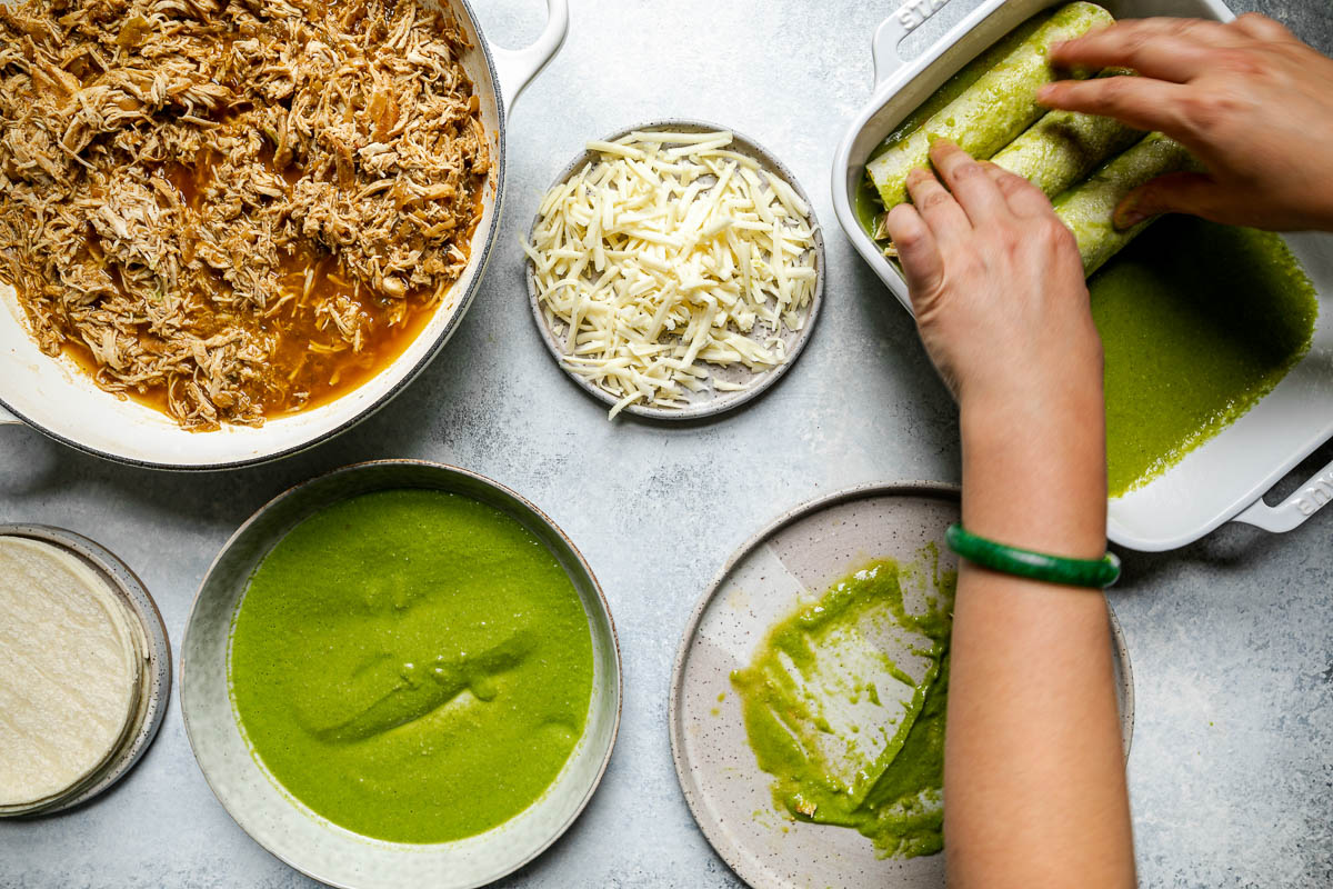 Green enchiladas assembly – green chicken enchiladas components (shredded chicken, shredded cheese, corn tortillas, & green enchiladas sauce atop a light blue surface. A woman's hands reach into the frame, placing an enchilada in a prepared baking dish with green enchilada sauce.