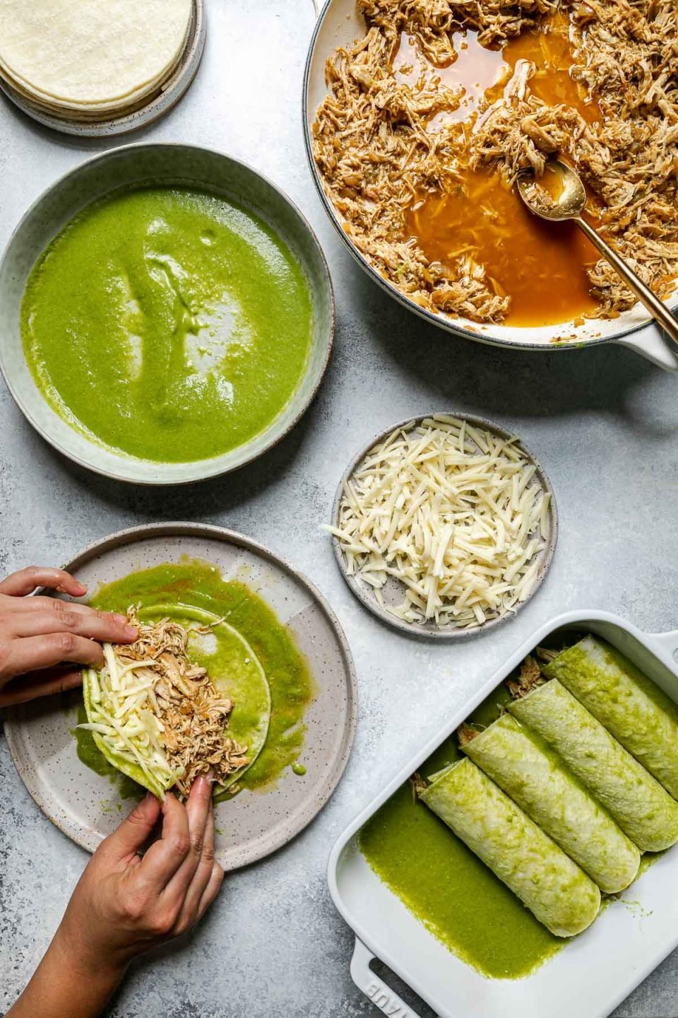 Green enchiladas assembly – green chicken enchiladas components (shredded chicken, shredded cheese, corn tortillas, & green enchiladas sauce atop a light blue surface. A woman's hands reach into the frame, wrapping chicken & cheese into a enchilada sauce-coated corn tortilla.