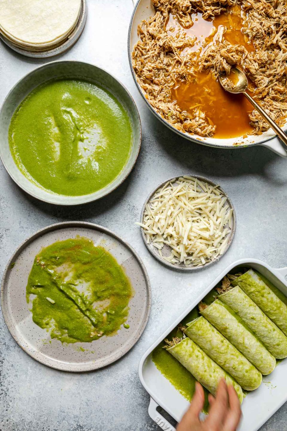 Green enchiladas assembly – green chicken enchiladas components (shredded chicken, shredded cheese, corn tortillas, & green enchiladas sauce atop a light blue surface. A woman's hands reach into the frame, placing an enchilada in a prepared baking dish with green enchilada sauce.
